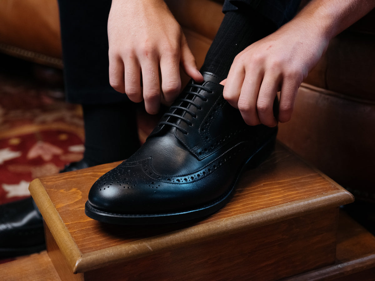 Wingtip Shoes in Raven Black, tying the laces with one foot on a wooden footstool