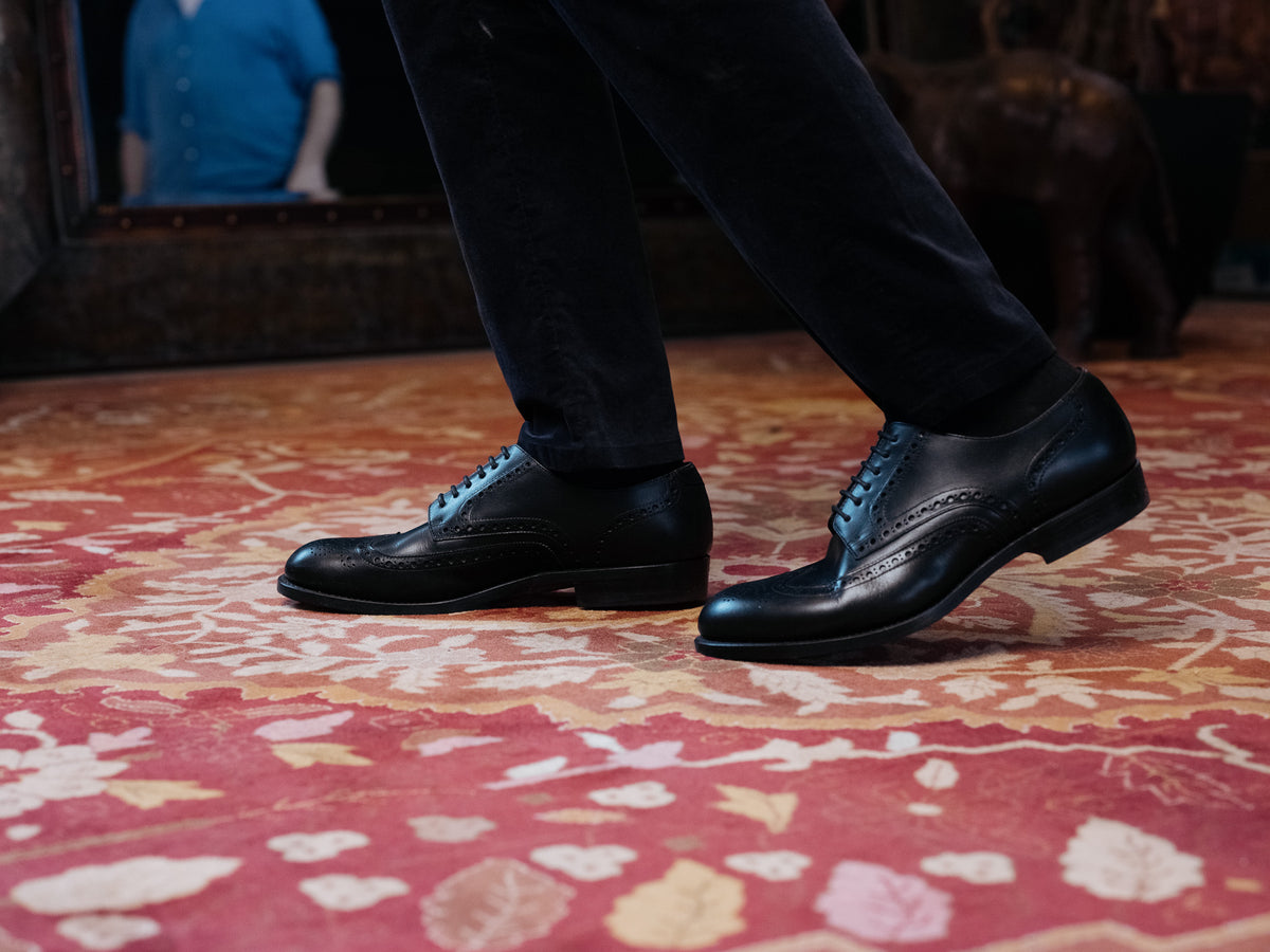 Wingtip Shoes in Raven Black, worn with dark trousers on a red patterned rug