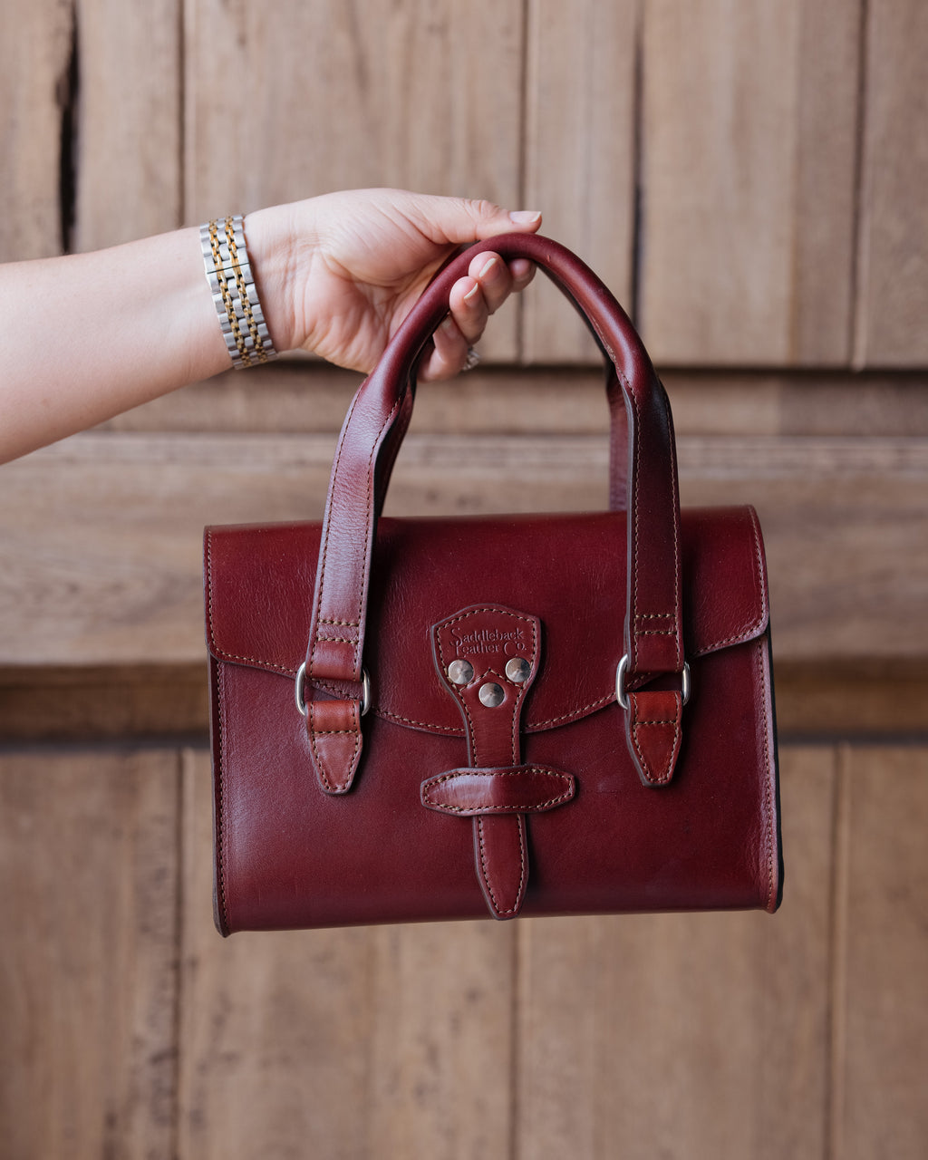 Double Handle Purse in Oxblood held up by its handles in front of a rustic wooden wall
