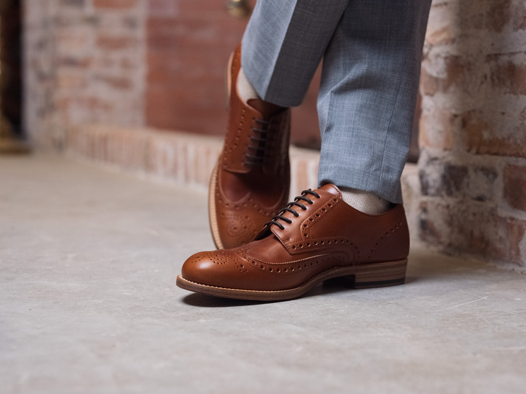 Wingtip Shoes in Natural Tan, worn mid-stride with gray trousers by a brick wall