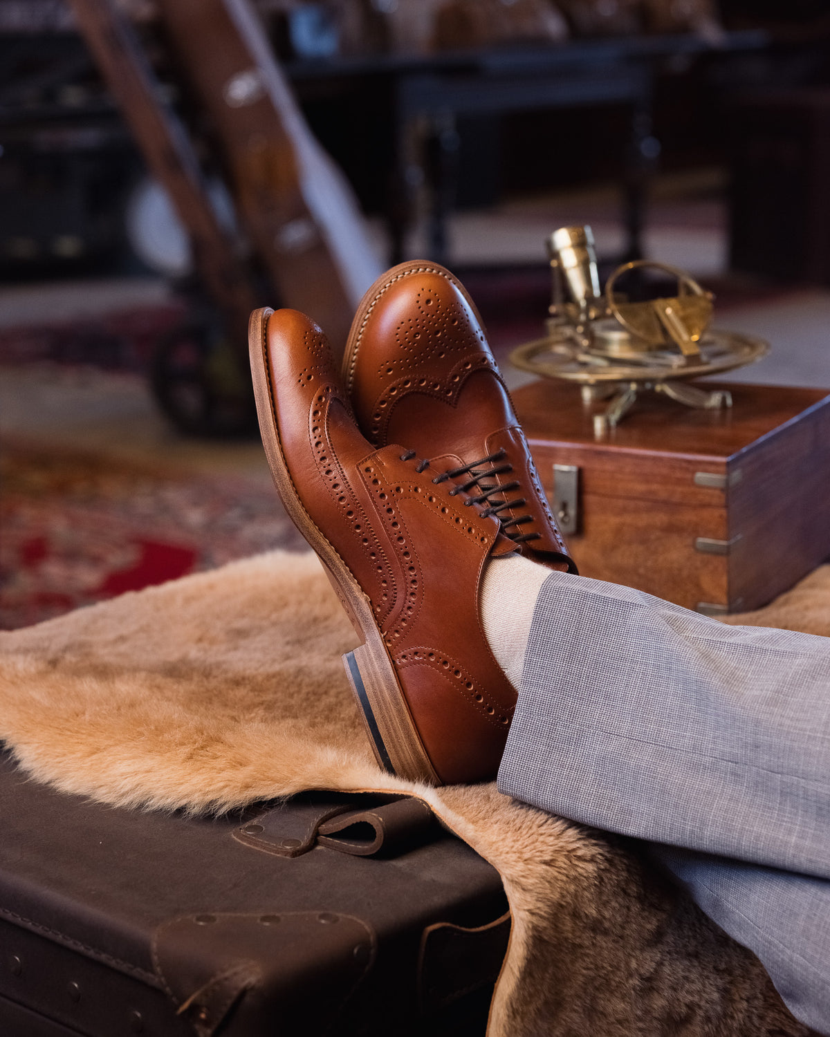 Wingtip Shoes in Natural Tan, crossed feet on a sheepskin beside a brass sextant and wooden box