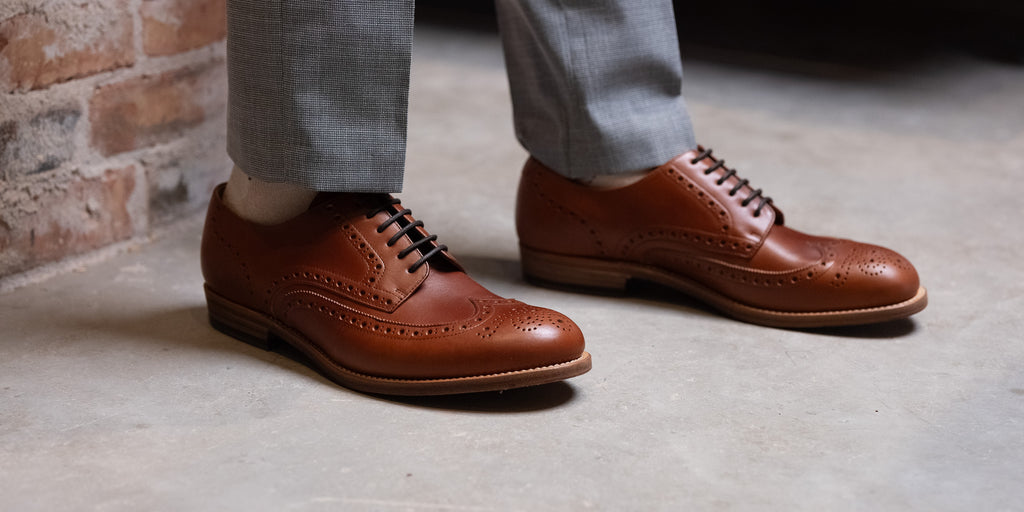 Wingtip Shoes in Natural Tan, low-angle view worn with gray trousers on concrete