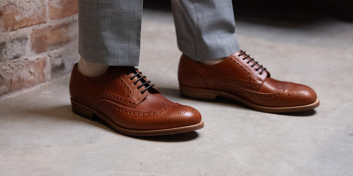 Wingtip Shoes in Natural Tan, low-angle view worn with gray trousers on concrete
