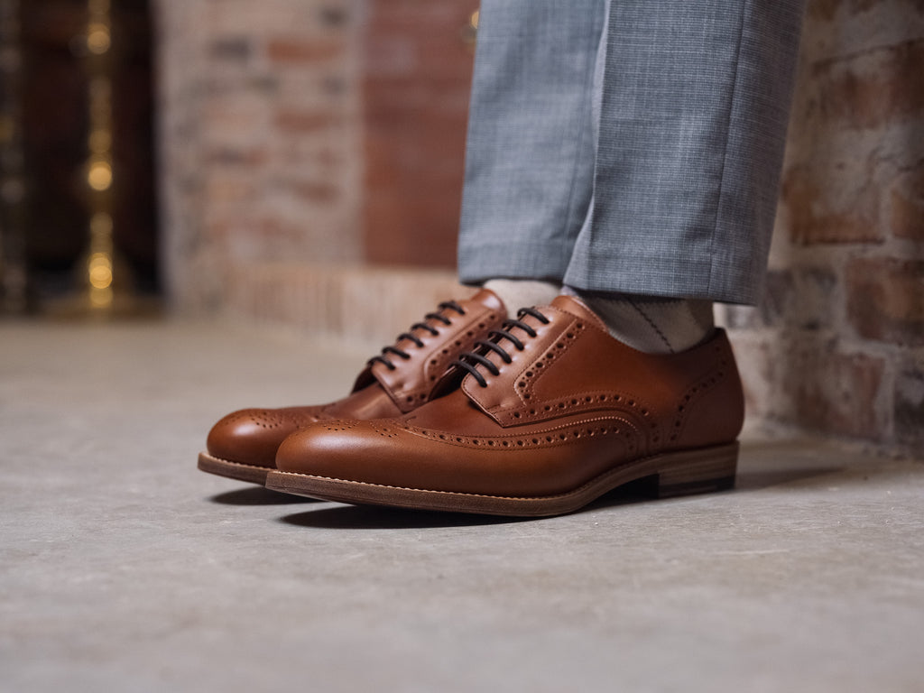 Wingtip Shoes in Natural Tan, worn with gray trousers on a concrete floor by a brick wall