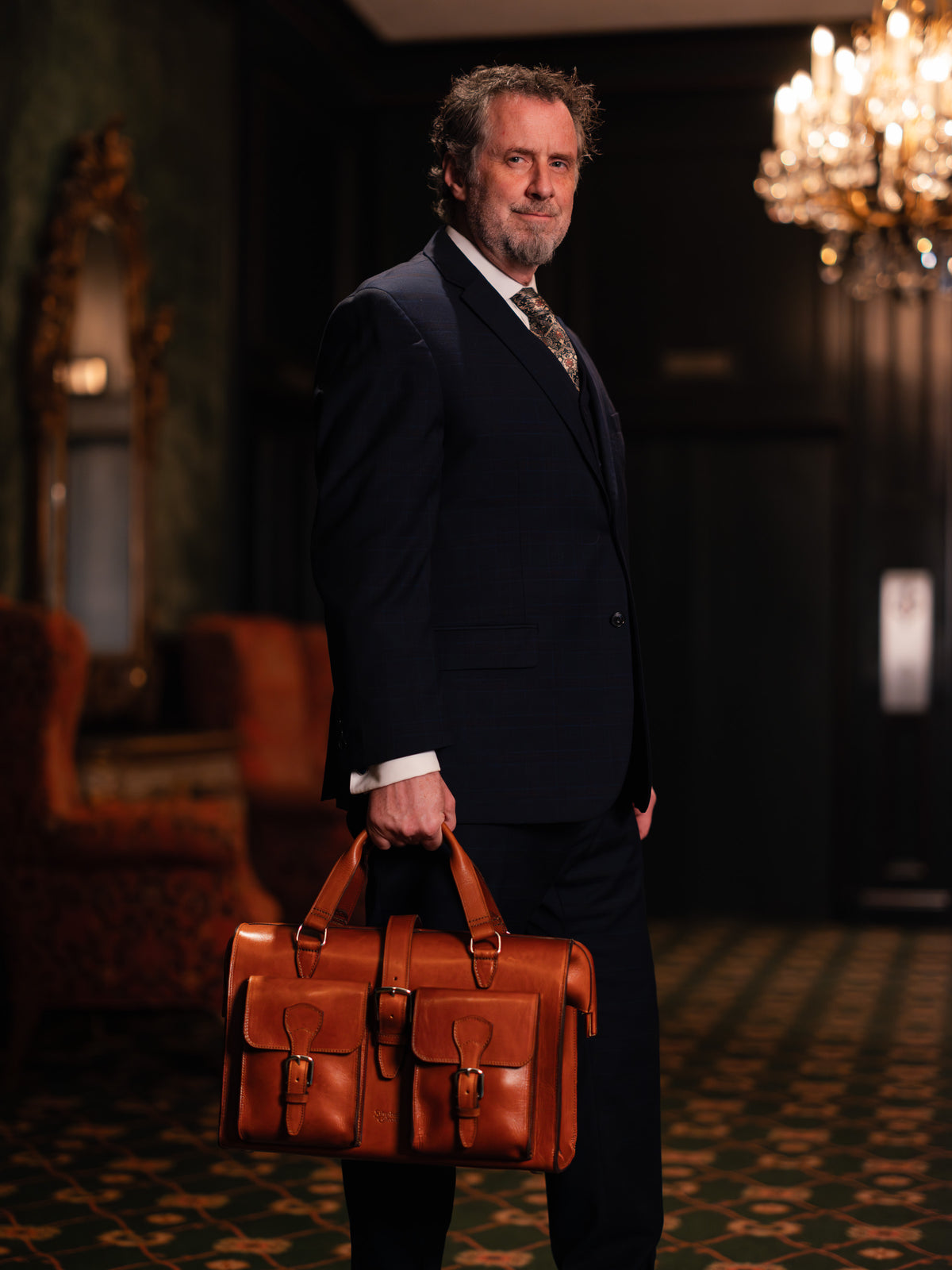 Blackstone Barrister Briefcase in English Tan carried by a man in a three-piece suit in a chandelier-lit room