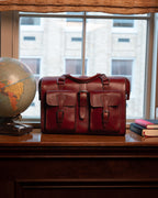 Blackstone Barrister Briefcase in Oxblood on a wooden sill by a window, between a globe and stacked books