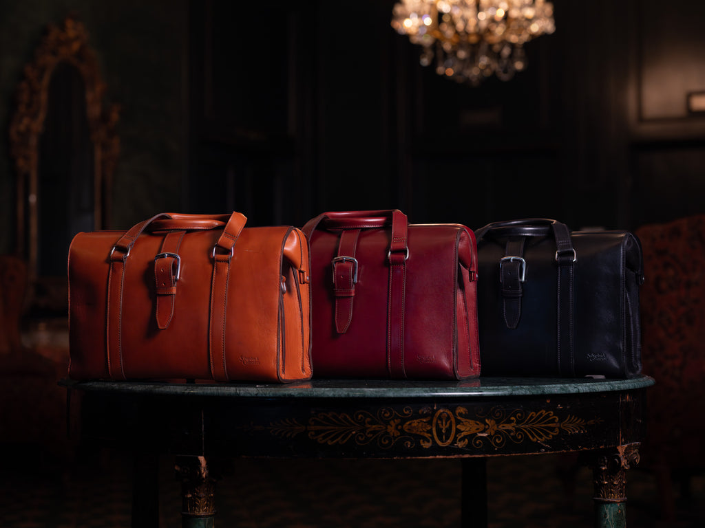 Spurgeon Briefcase in English Tan, Oxblood and Raven Black side by side on a marble table under a chandelier