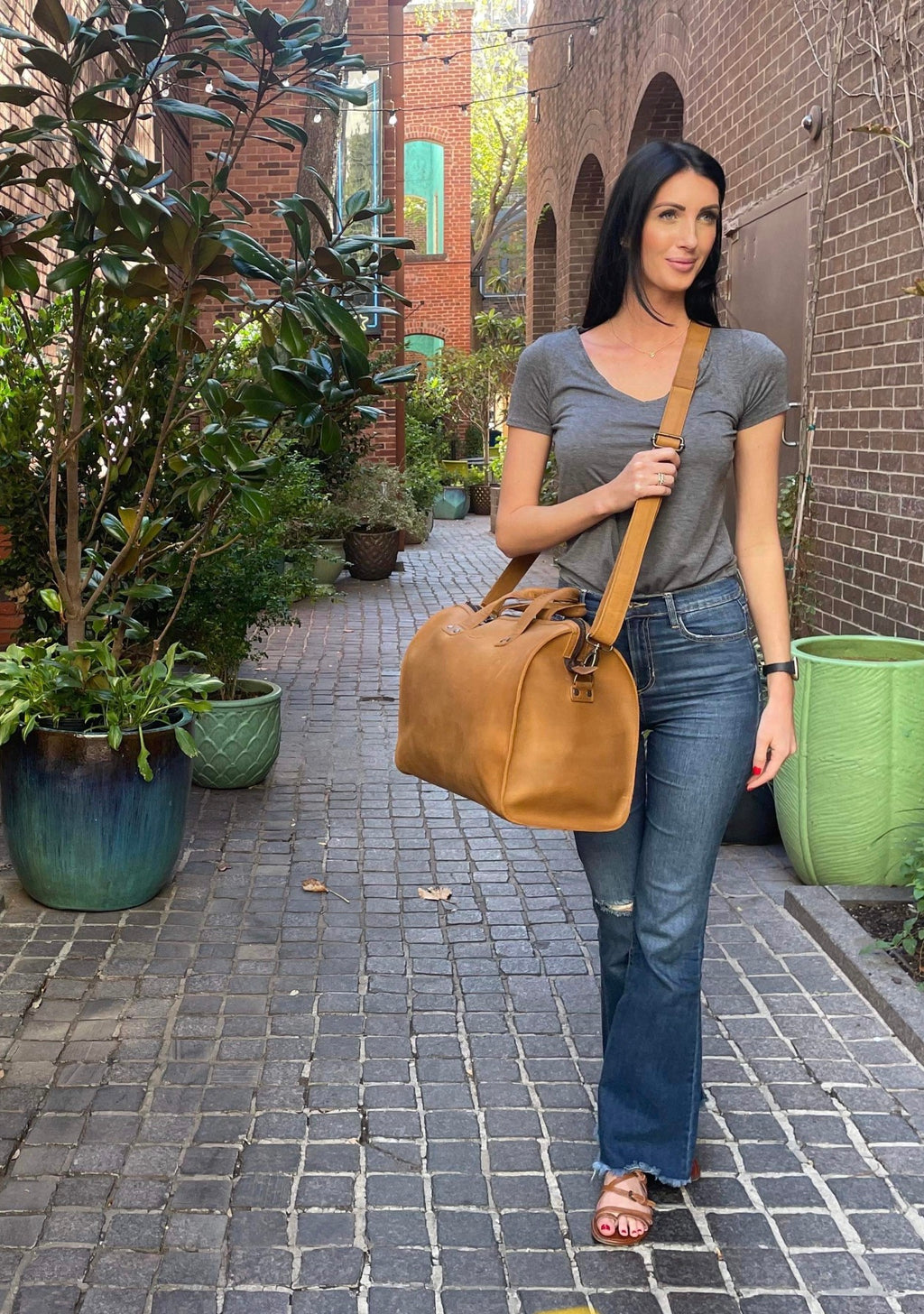 Simple Overnight Leather Duffle Bag in Tobacco, worn crossbody by a woman standing in a brick courtyard