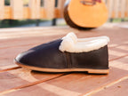 Sheepskin Moccasin in Dark Coffee Brown with shearling cuff, side view on a wooden deck with a guitar behind