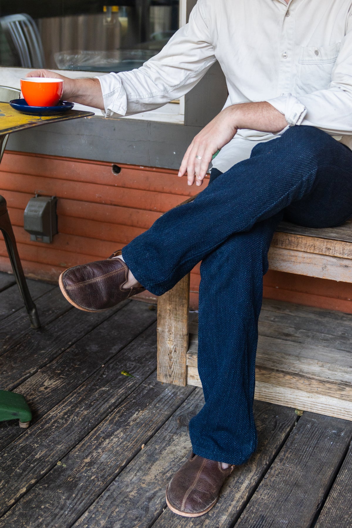 A man with a coffee cup seated on a wooden porch bench wearing Barefoot Slip-on Shoes in Dark Coffee Brown