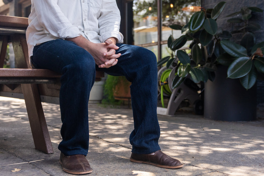 A man in jeans seated on a patio bench wearing Barefoot Slip-on Shoes in Dark Coffee Brown