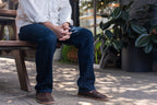 A man in jeans seated on a patio bench wearing Barefoot Slip-on Shoes in Dark Coffee Brown