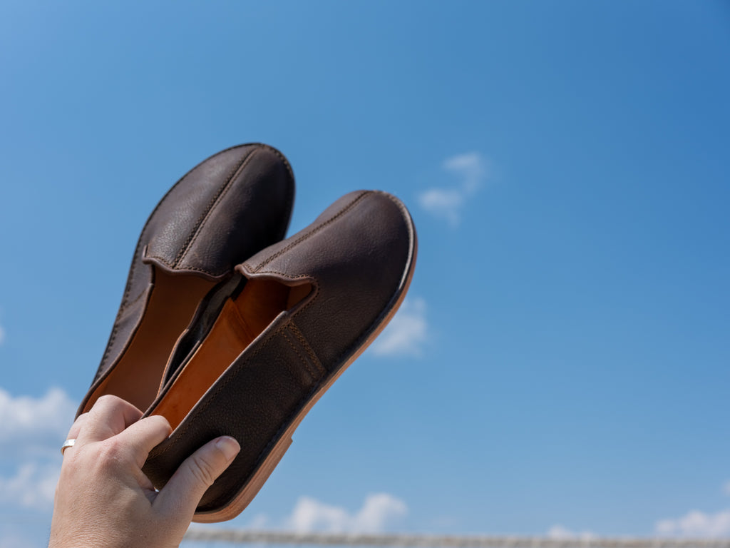 A hand holding up a pair of Barefoot Slip-on Shoes in Dark Coffee Brown against a blue sky