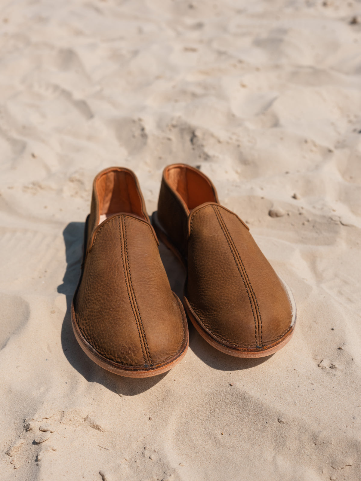 Pair of Barefoot Slip-on Shoes in Tobacco leather standing on beach sand, front view