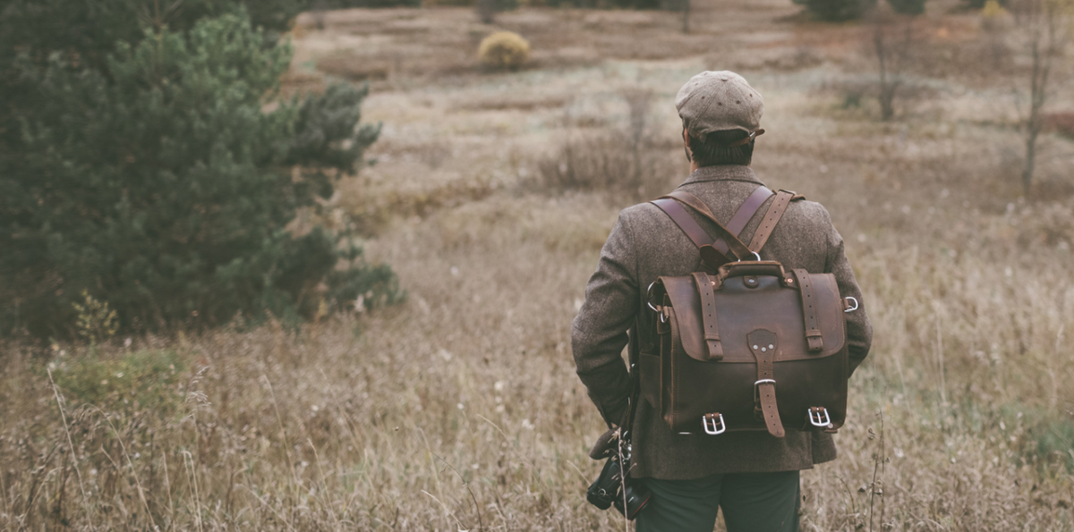 A man in a tweed cap and jacket, wearing a large leather backpack, looks out over a field of tall grass and pine trees in autumn.