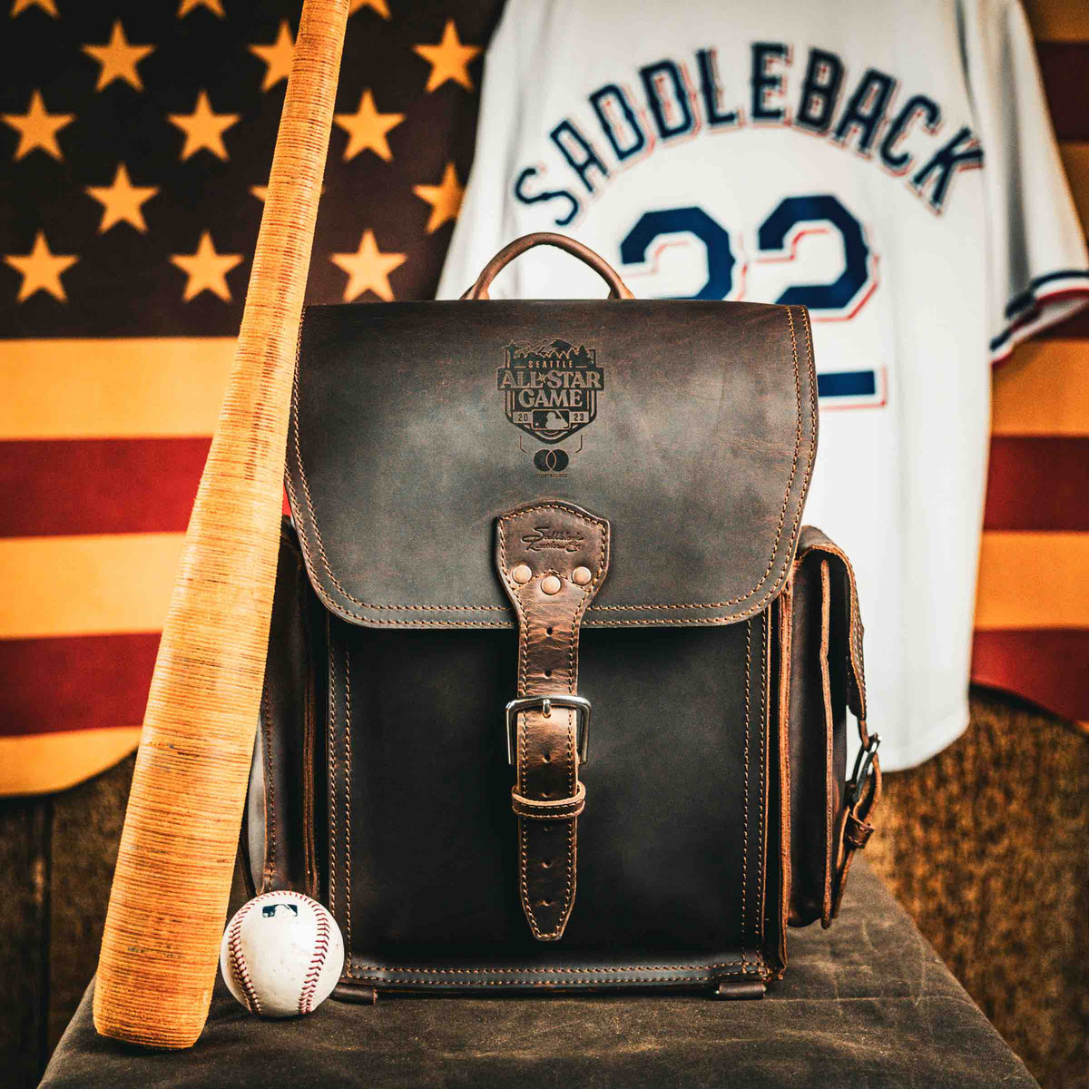 A dark brown Tank Leather Backpack stamped with the 2023 All-Star Game logo, beside a bat and a baseball, in front of a Saddleback number 22 jersey and an American flag.