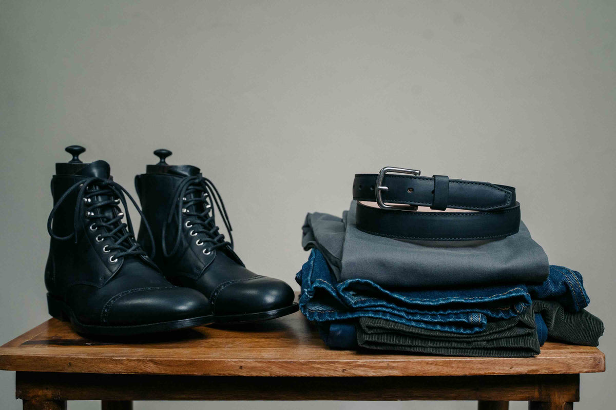 Italian Dress Belt in Raven Black, coiled on folded jeans beside black leather boots