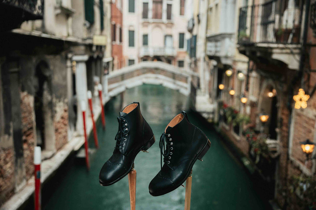 Pair of high quality black leather Officer Boots on wooden stands above a Venice canal, side view