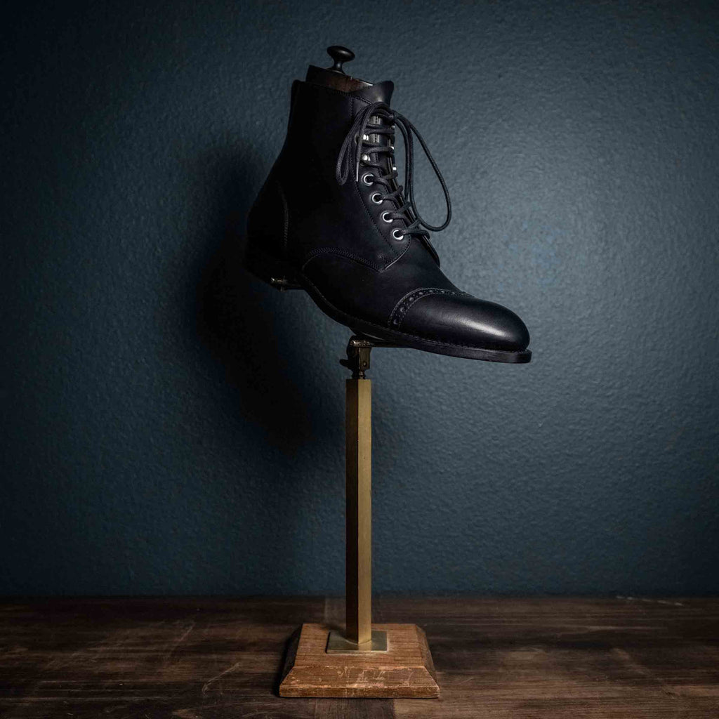 Single high quality black leather Officer Boot on a brass display stand, side view against a dark wall