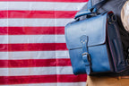 Saddleback Club Membership Paul Revere Leather Messenger Bag Backpack in Red, White and Blue, worn in front of an American flag