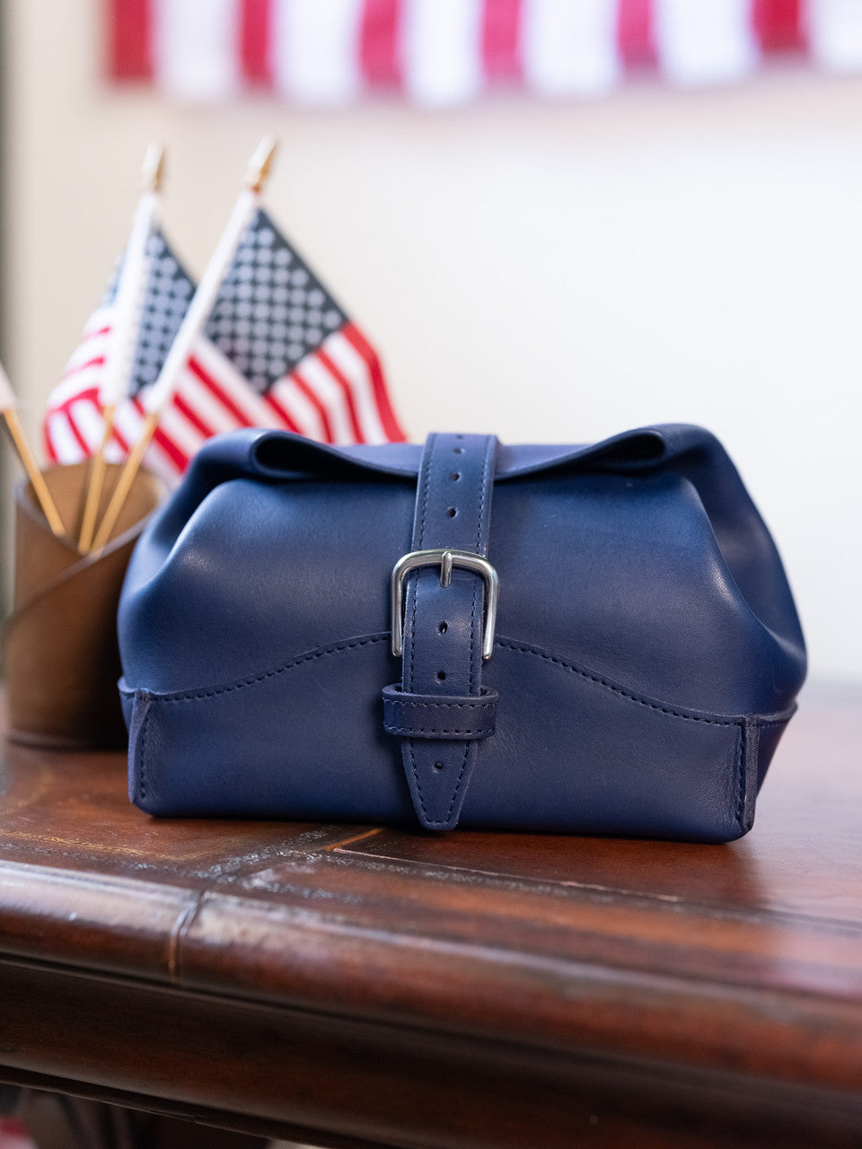 Saddleback Club Membership George Washington Leather Roll-Up Toiletry Bag in Blue on a wood desk with flags and bunting behind