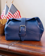 Saddleback Club Membership George Washington Leather Roll-Up Toiletry Bag in Blue, front view on a desk with flags behind