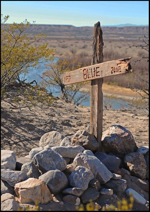 A rough wooden cross carved BLUE, with the years 1997 and 2008, standing in a pile of stones on a desert hillside above a river.