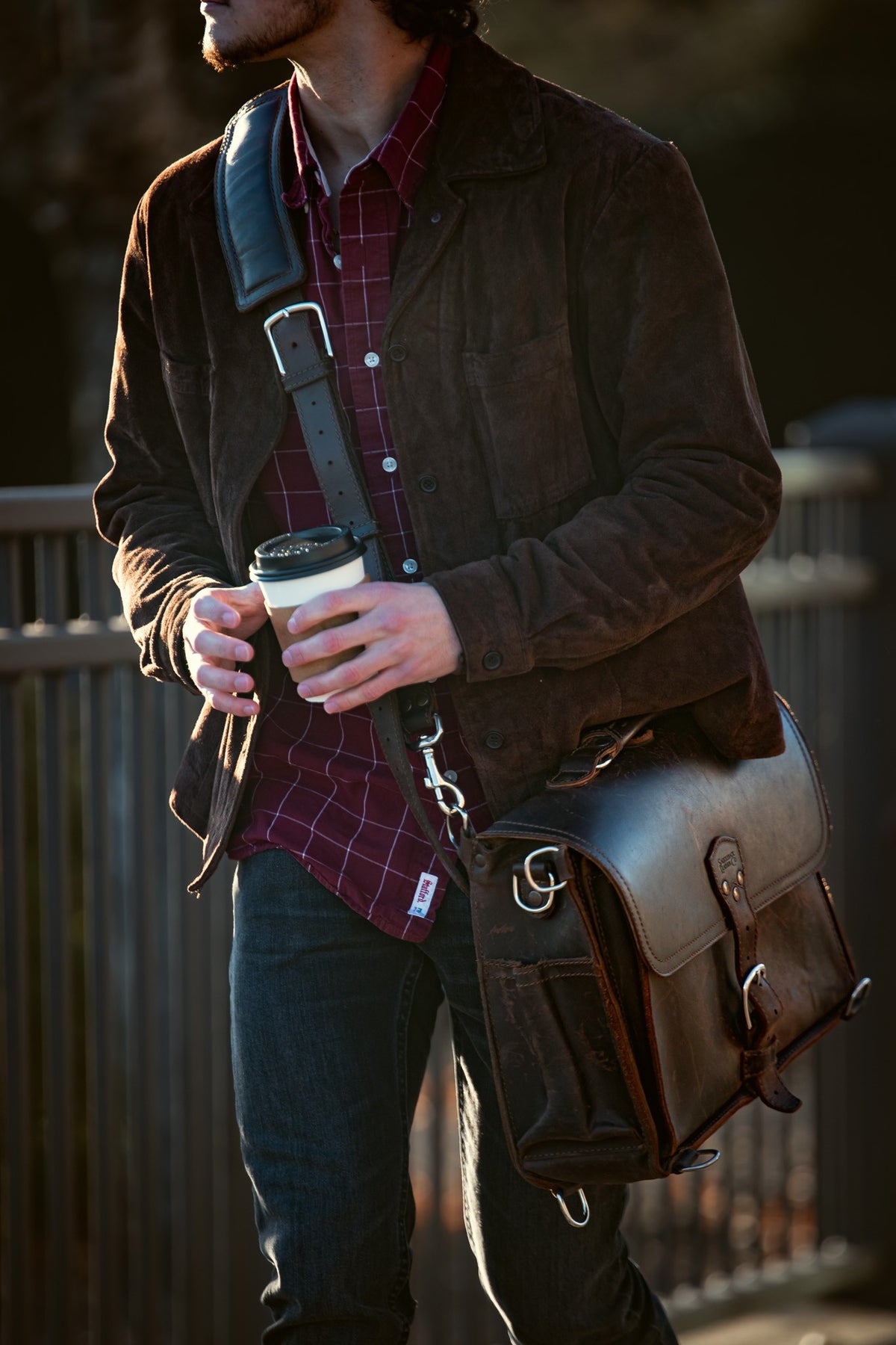 Man in a brown jacket holding a coffee cup, carrying a dark brown leather slim briefcase on a shoulder strap