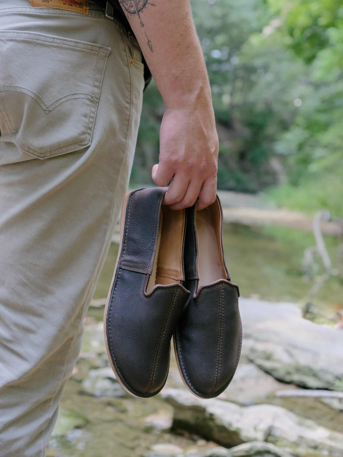 Barefoot Slip-on Shoe in Dark Coffee Brown, pair held in a hand beside a rocky river