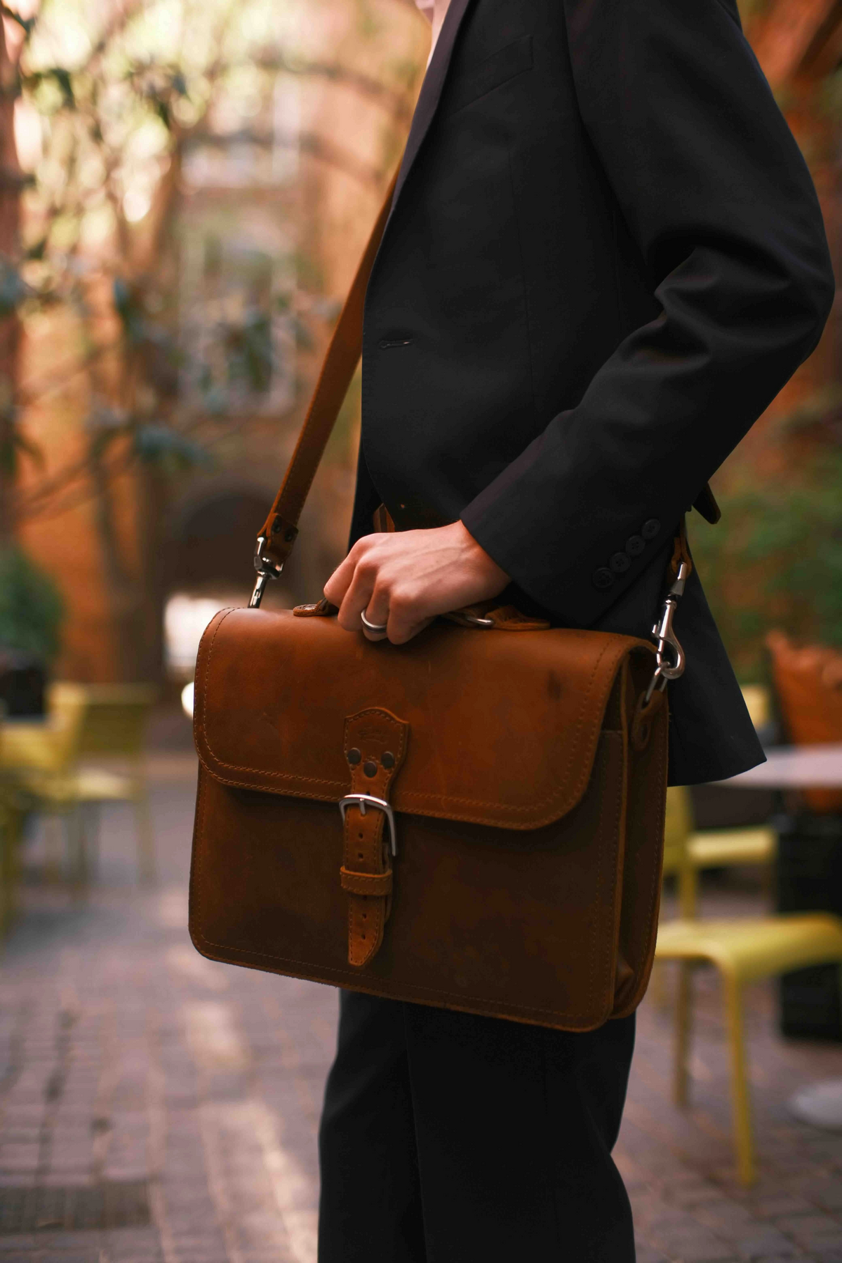 A person in a black blazer and trousers carries a brown leather briefcase with a buckle strap in an outdoor patio setting.