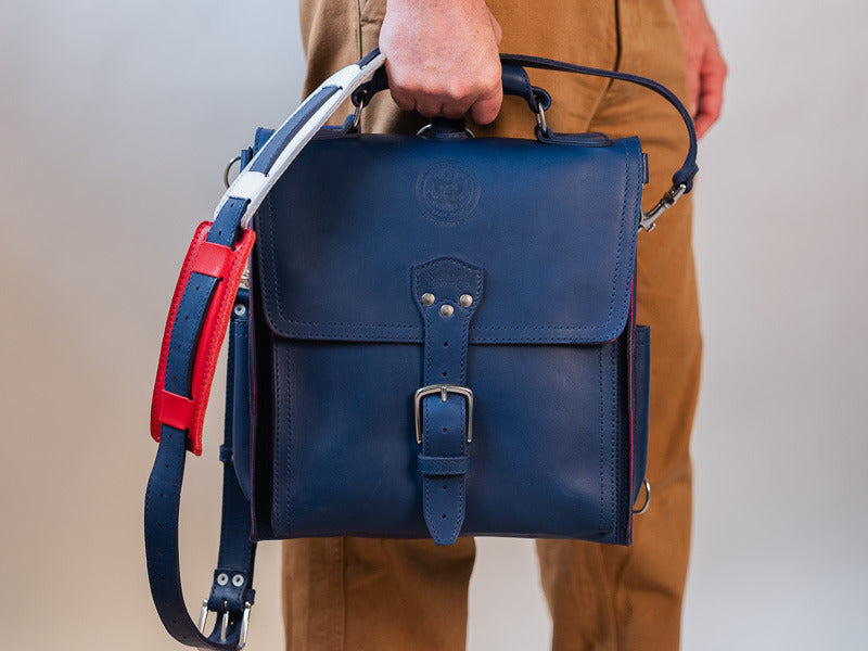 A man carrying the navy messenger bag by its top handle, its red and white strap alongside