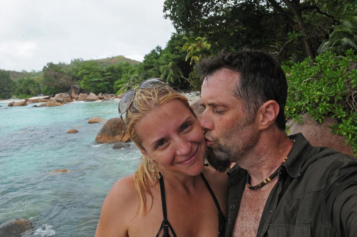 A man kissing the cheek of a smiling blonde woman in a selfie on a tropical beach with turquoise water and granite boulders.