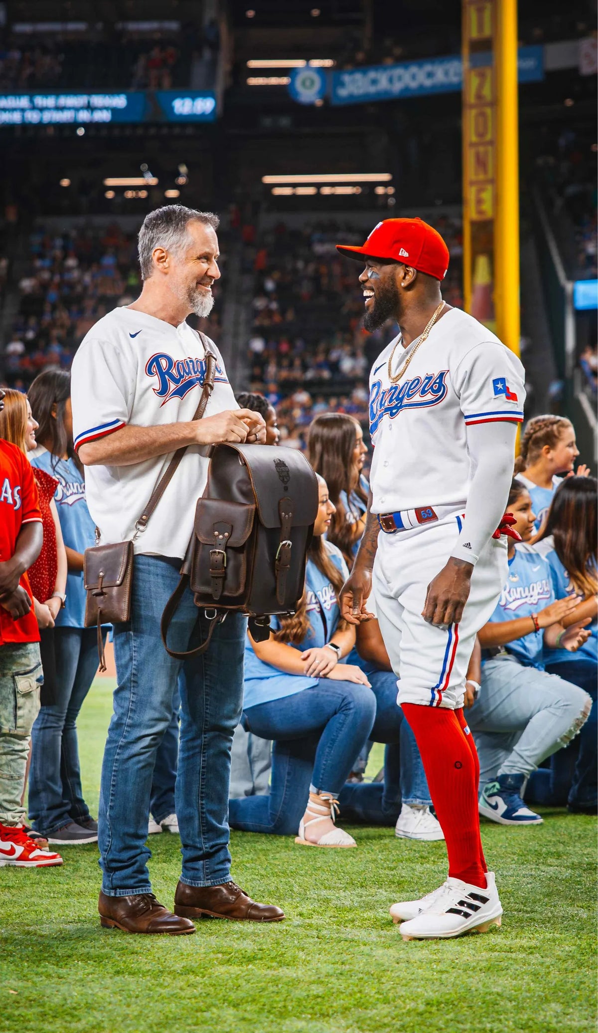 Dave Munson, in a Rangers jersey, talking with Adolis Garcia on the field as he presents him with a dark brown Tank Leather Backpack, fans in the stands behind.