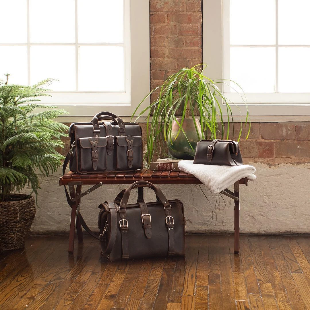 Three dark brown leather bags, including a Doctor's Duffle, on and beside a rustic bench in a brick-walled room with plants