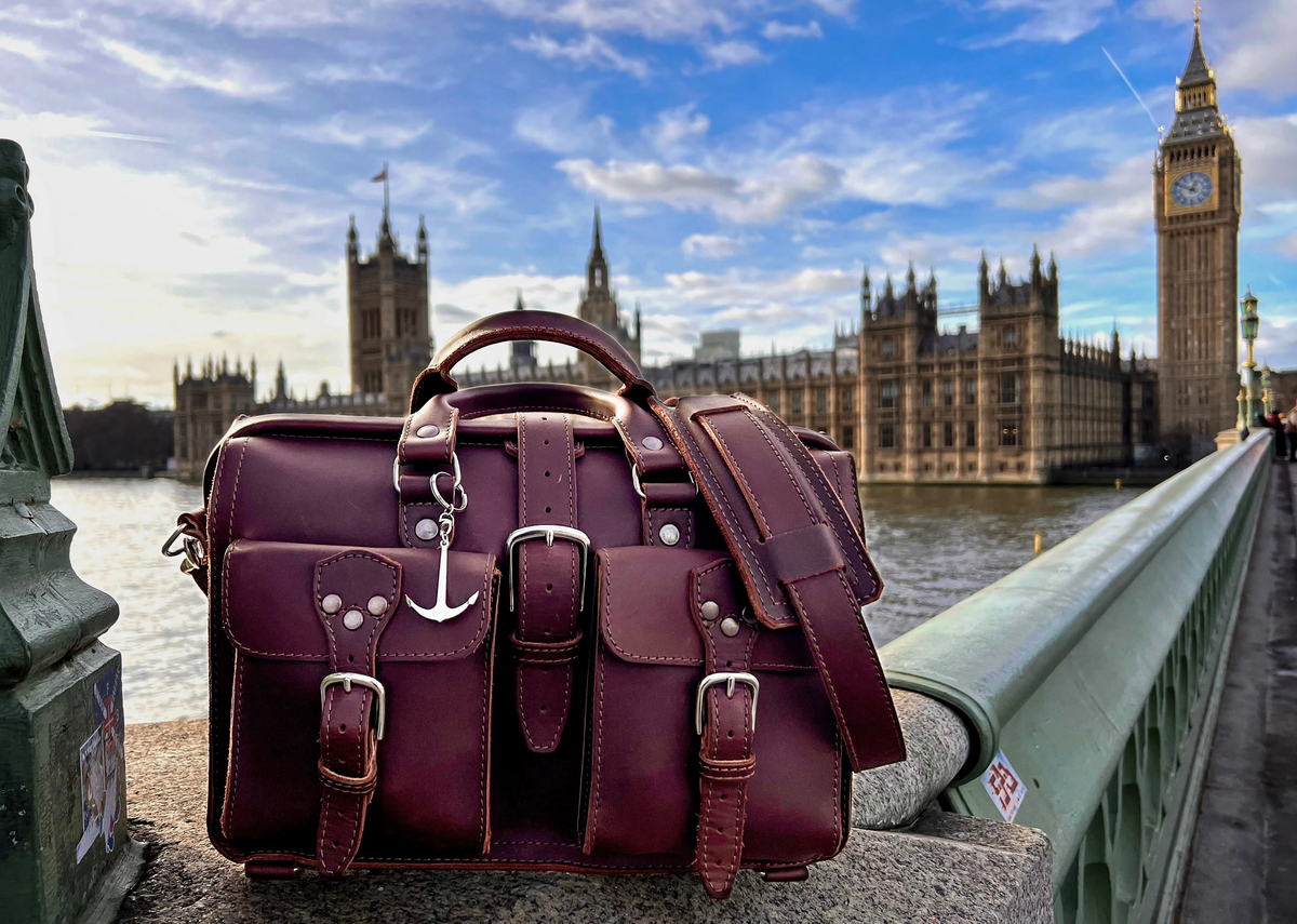 Burgundy leather Flight Bag briefcase with an anchor charm on a bridge ledge in front of Big Ben in London