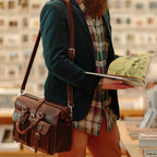 Man in a bookshop reading a book while carrying the Flight Bag Leather Briefcase in chestnut on its shoulder strap