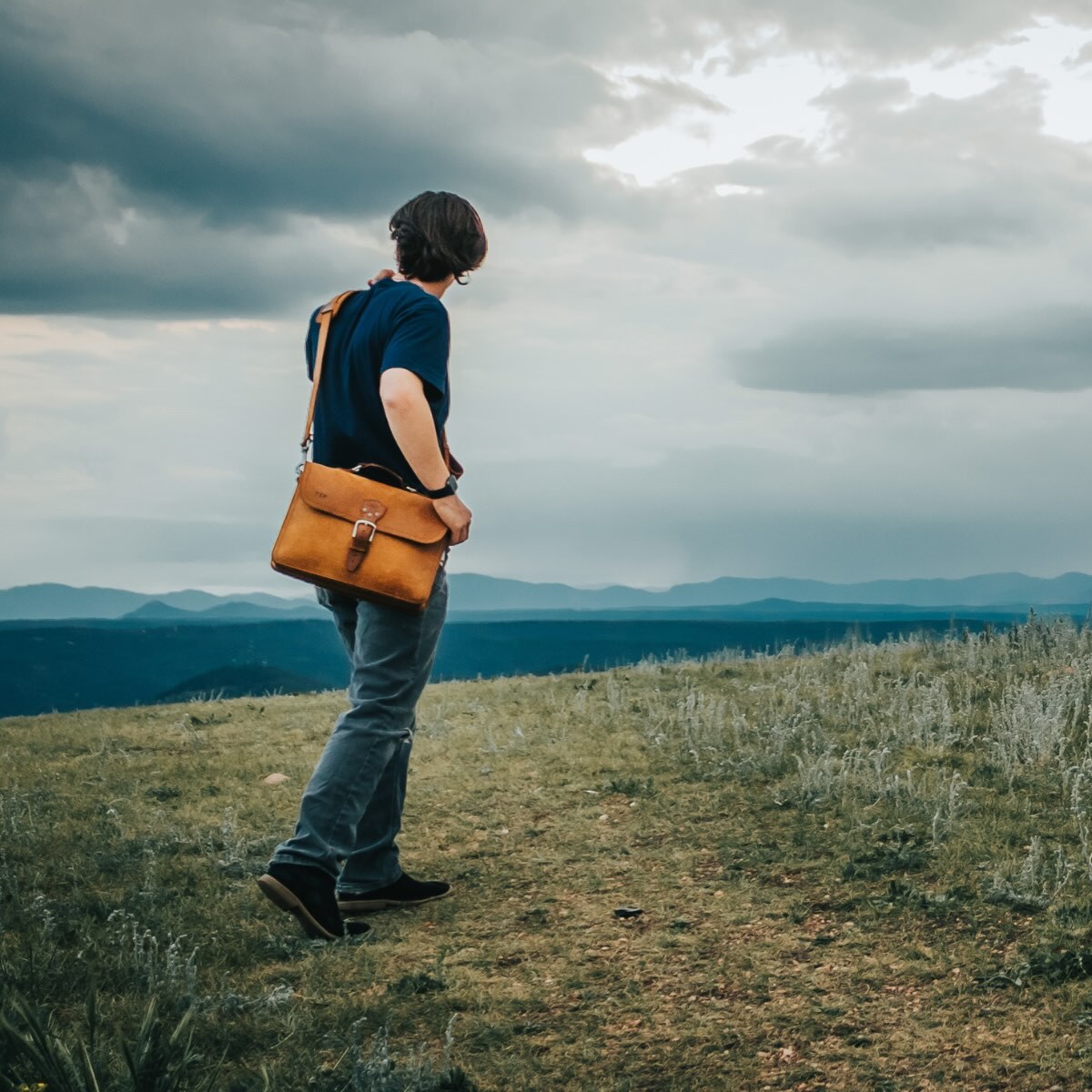 Man on a grassy hilltop overlooking mountains, carrying a tan Fourteener thin leather briefcase over his shoulder