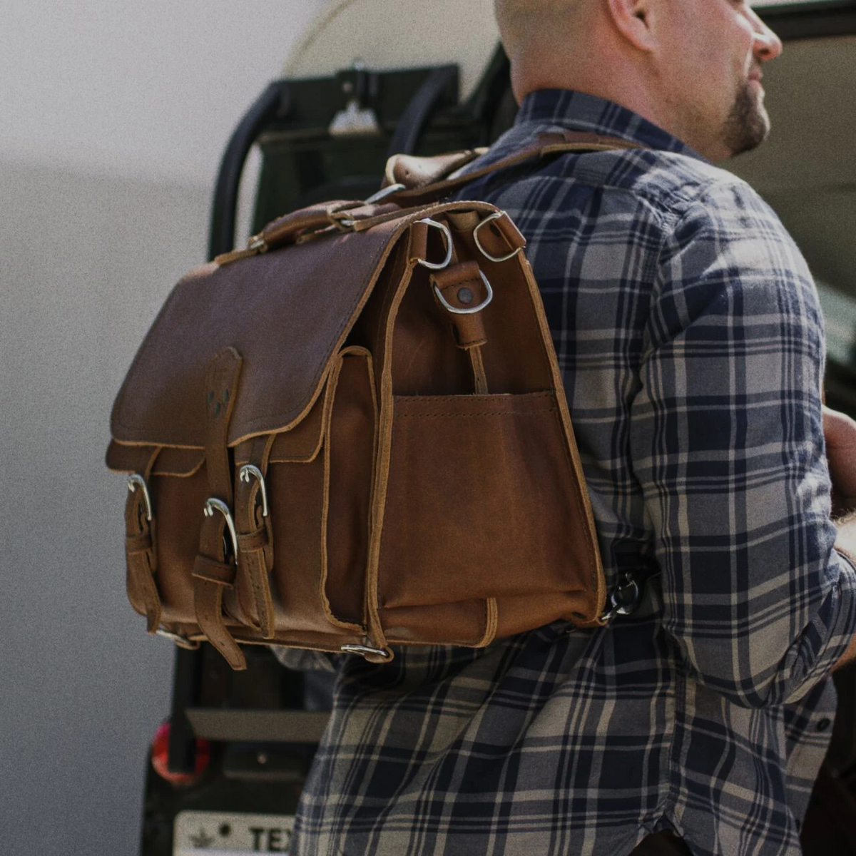 Man in a plaid shirt wearing a large chestnut leather front pocket briefcase as a backpack