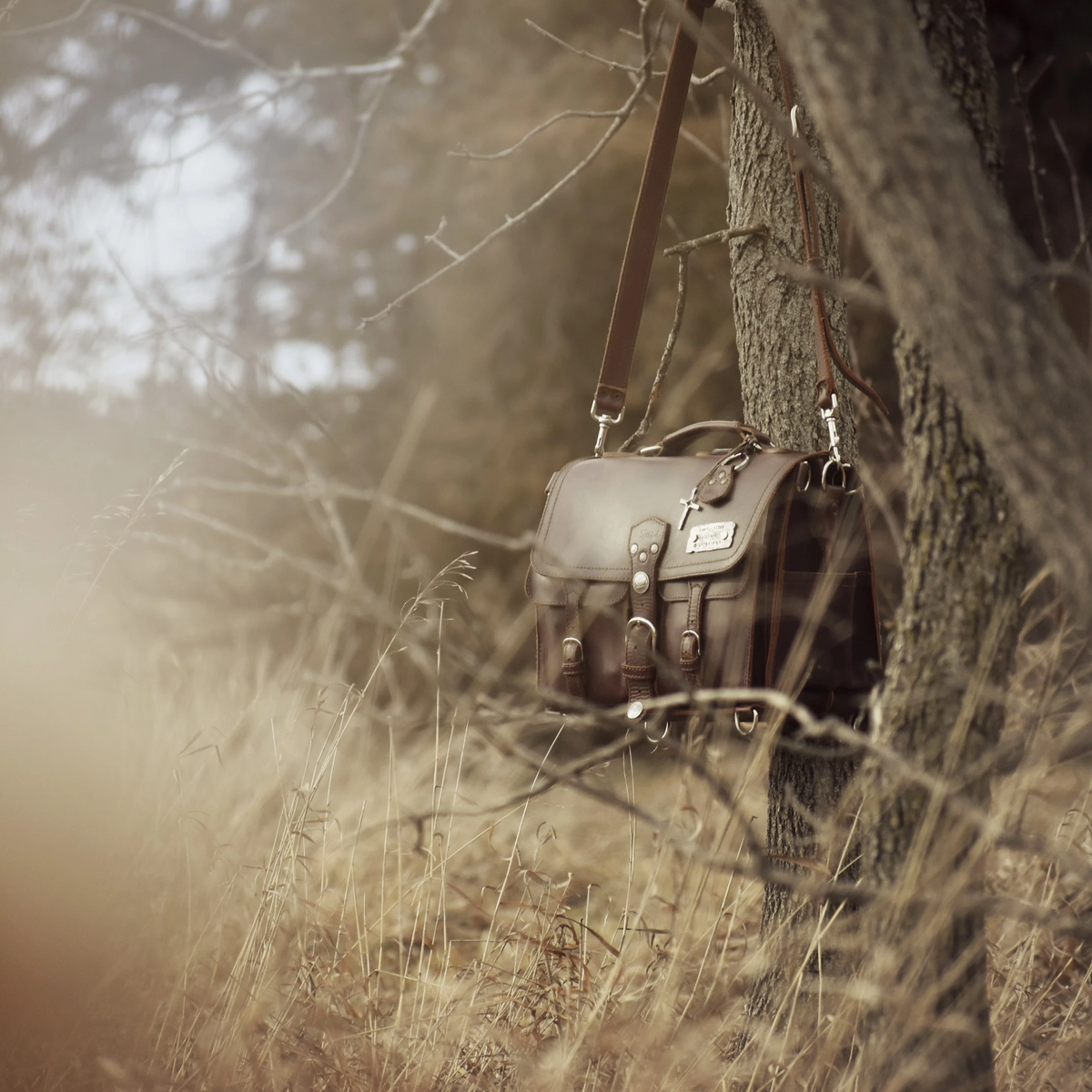 Saddleback Leather front pocket briefcase hanging by its shoulder strap from a tree in dry tall grass