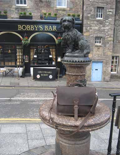 The bronze statue of Greyfriars Bobby on its granite fountain in Edinburgh, in front of Bobby's Bar, with a dark brown leather satchel set on the fountain's ledge.
