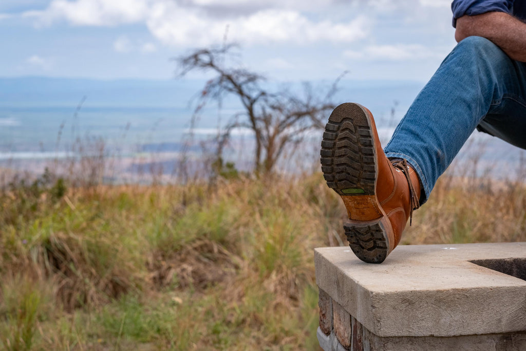 Huntsman Boots in brown leather, raised foot showing lug sole while seated on a stone ledge outdoors