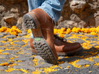 Huntsman Boots in brown leather, worn with jeans mid-step on a stone path scattered with marigold petals