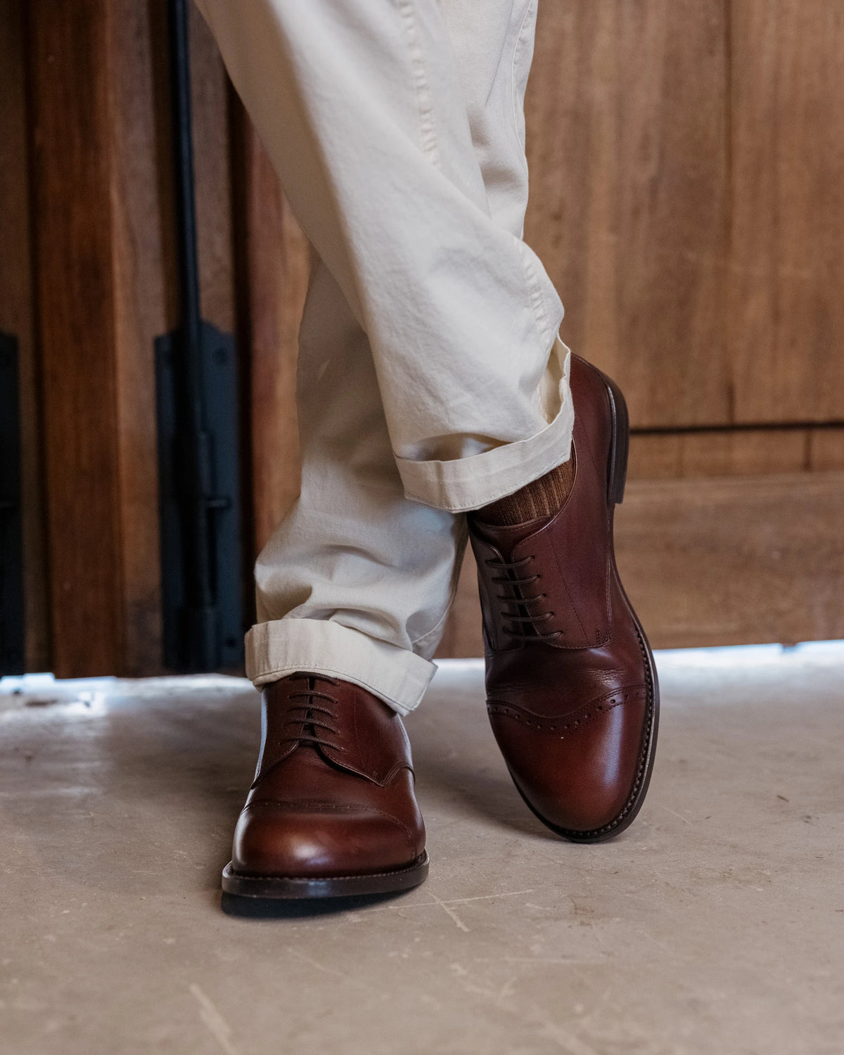 Toe Cap Shoes - Dark Oak worn with crossed legs and cuffed cream trousers before a wooden door
