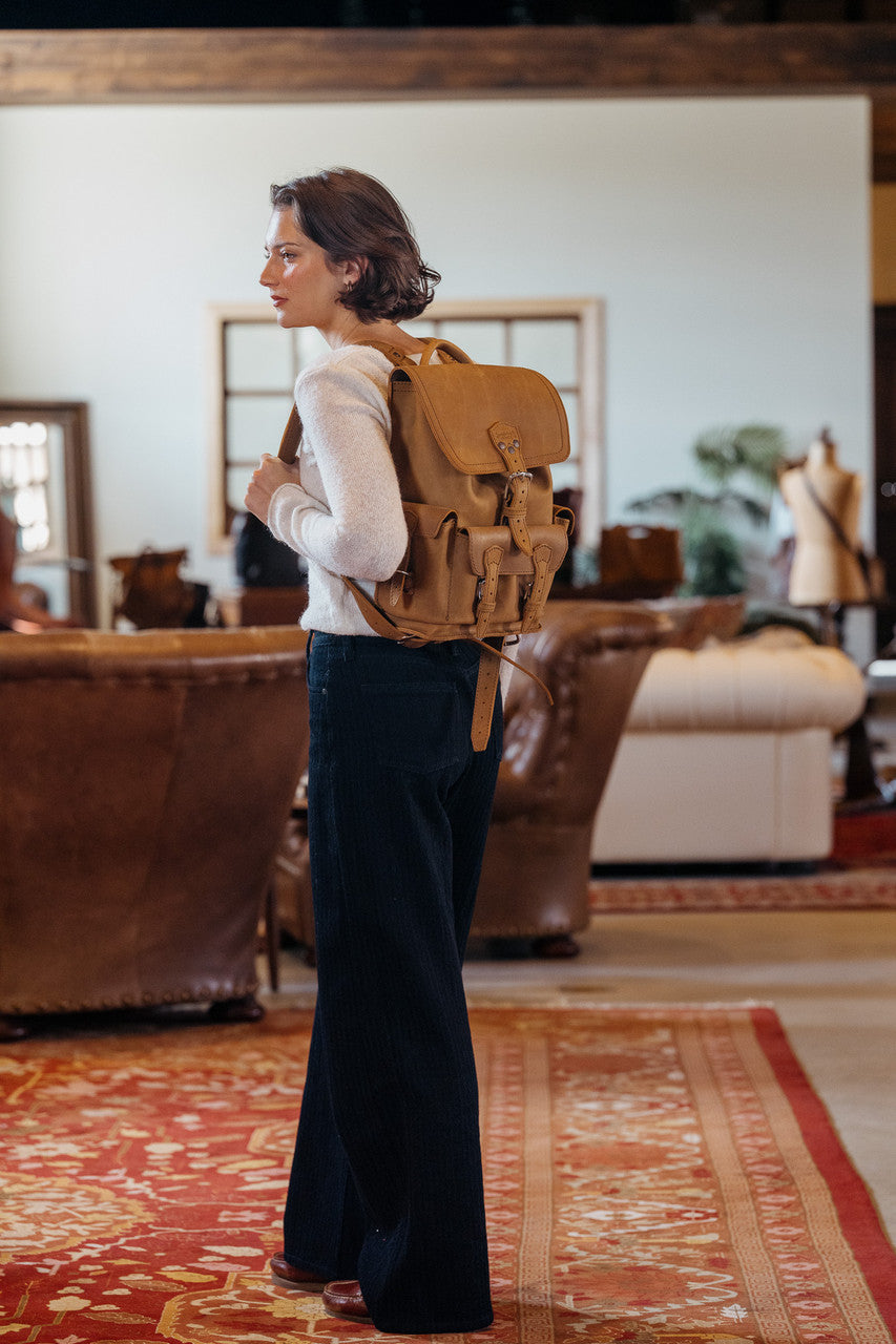 Small Front Pocket Backpack in Tobacco worn by a woman in profile in a rustic room with leather sofas and a red rug