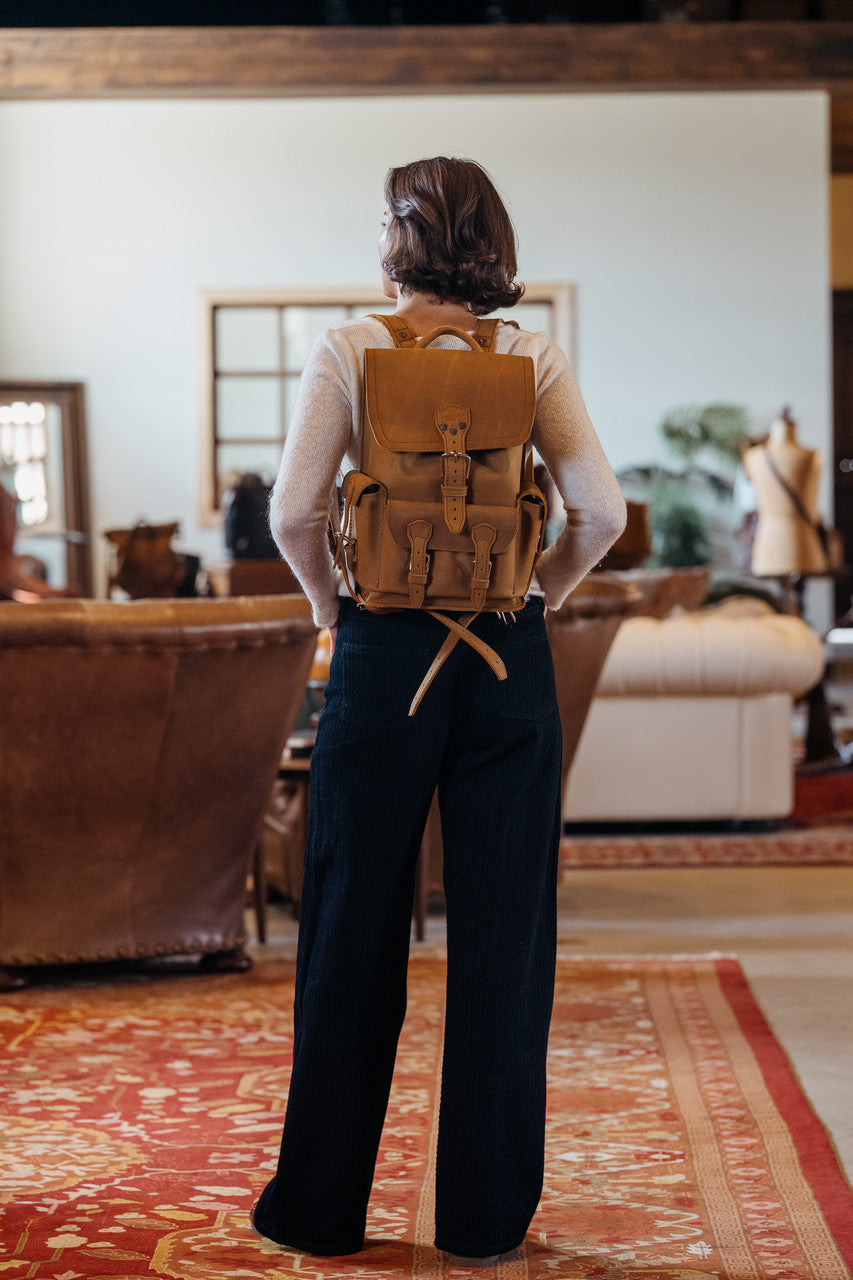 Small Front Pocket Backpack in Tobacco worn by a woman seen from behind in a rustic room with leather sofas and a red rug