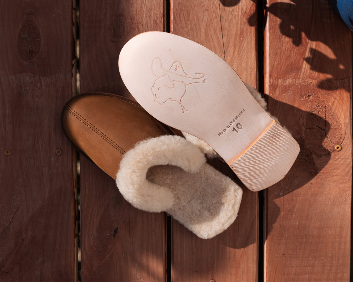 Men's Sheepskin Slippers in Tobacco, one slipper beside an upturned sole with stamped dog logo on a deck
