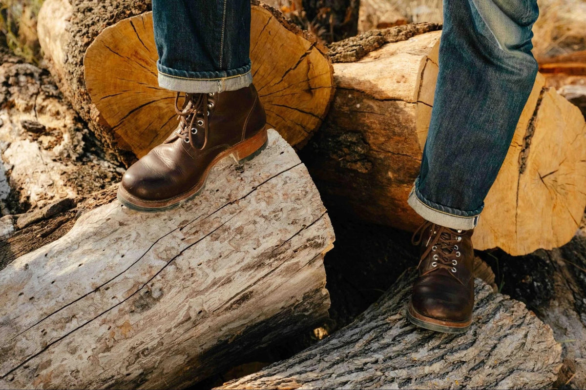 Huntsman Boots - Dark Coffee Brown on foot with cuffed jeans, climbing over a pile of cut logs