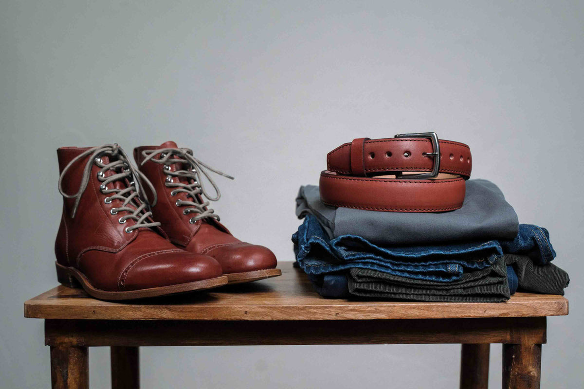 Officer Boot in Oxblood, pair on a wooden table beside a matching belt and folded jeans