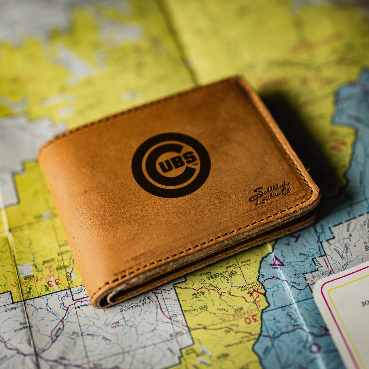 Chicago Cubs Leather Bifold Wallet in Tobacco resting on a colorful road map, close-up of the logo