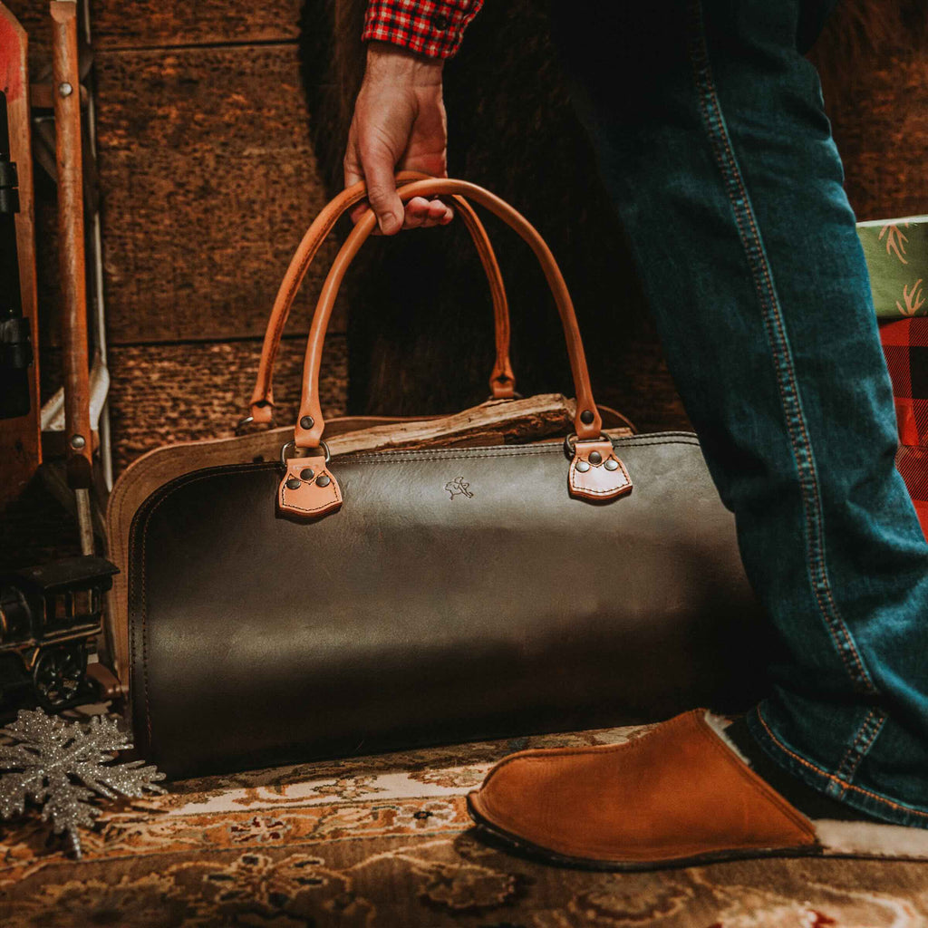 Man in jeans and slippers lifting the Leather Log Carrier in Dark Coffee Brown by its handles on a rug